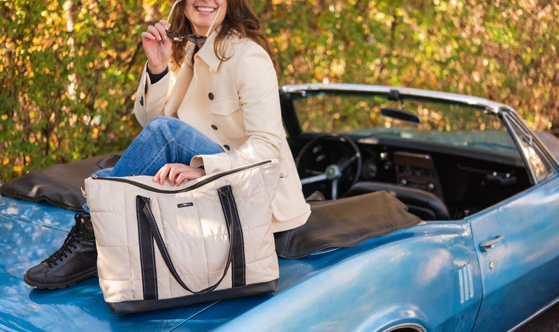 A woman sitting on the hood of a blue convertible car holds a large cream quilted tote bag with black handles and trim. The background shows a blurred outdoor setting with autumn foliage.
