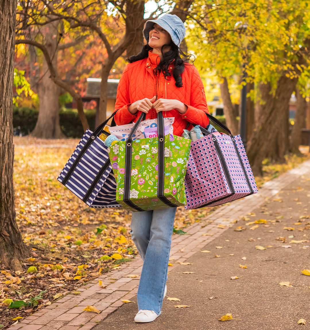 A woman walking on an autumn park path carrying three colorful shopping bags: a navy and white striped bag, a green floral bag, and a pink polka dot bag.